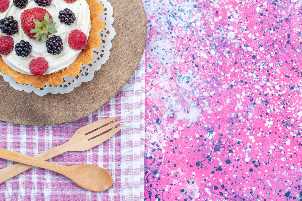 Berry-topped cake on a wooden board with a lace doily, on a pink plaid cloth and wooden cutlery, against a pink paint-splatter background.