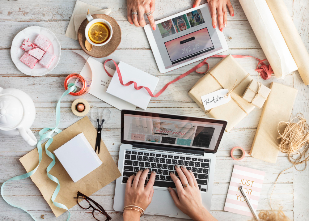 Overhead view of a desk with gift boxes, ribbons, a laptop, tablet, tea, and cookies.