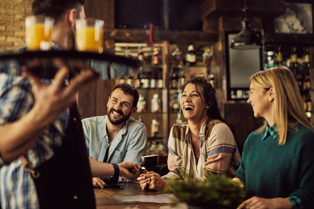 Three friends sit at a bar, smiling and chatting as a bartender brings a tray of drinks toward them .