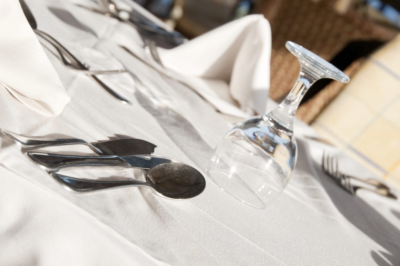 Elegant table setting with silverware laid out on a white tablecloth and a tilted empty wine glass nearby, outdoors on a sunny day.