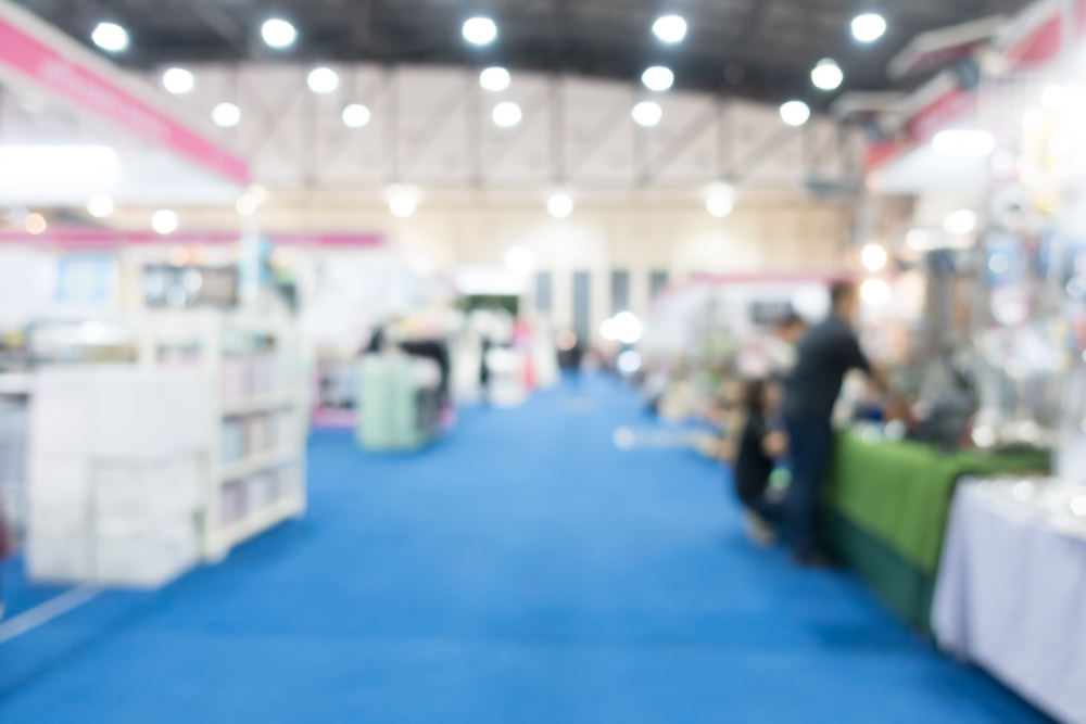 Blurred view of an indoor trade show with blue aisle carpet and booths on both sides under bright overhead lights.