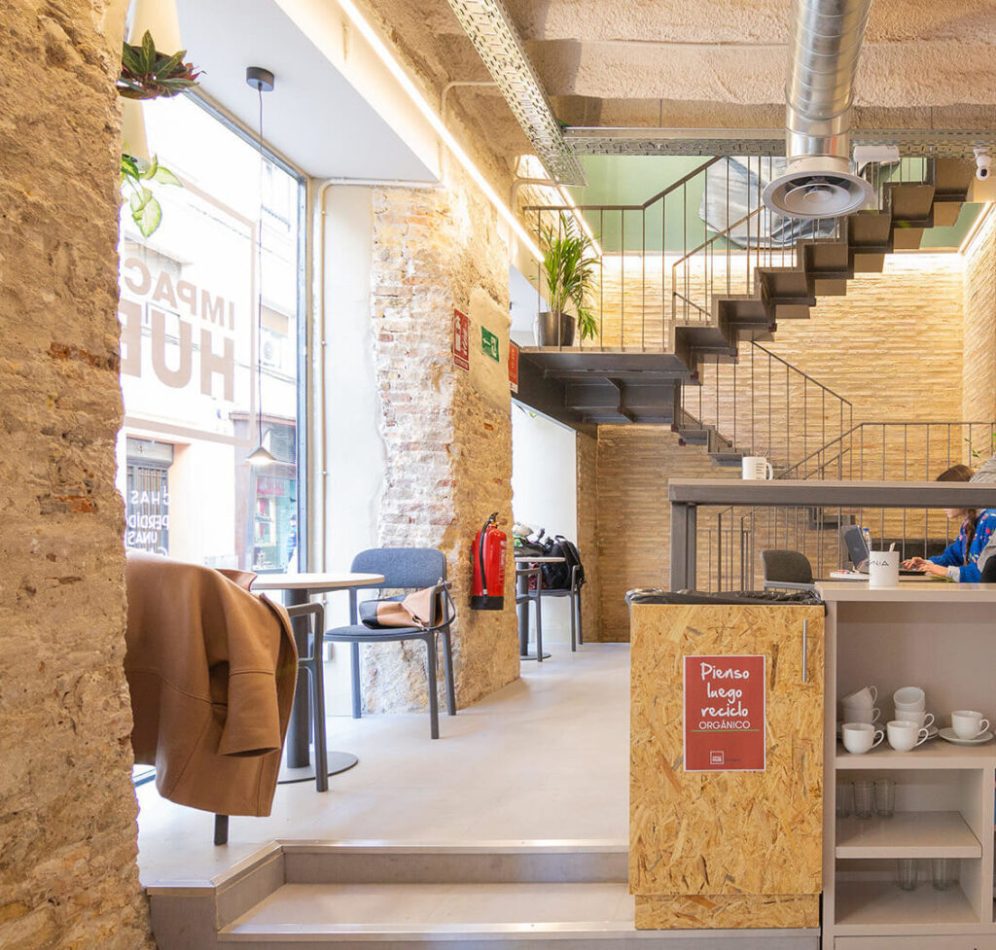 Bright cafe interior with brick walls, a metal staircase, and a counter with cups; a brown jacket draped over a chair. | PymesAutonomos.com