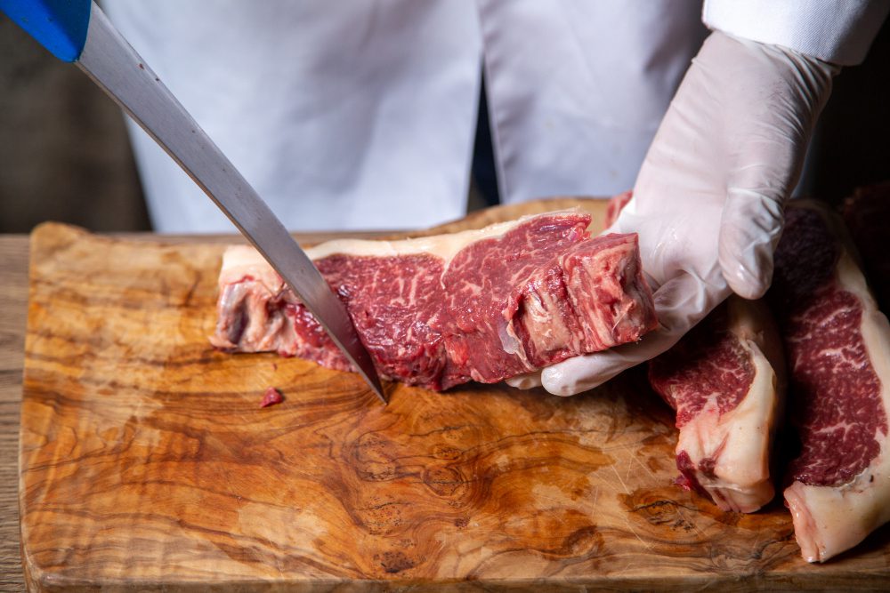 780972 - Menceyes Food Company | PymesAutonomos.com Butcher wearing gloves slices raw beef with a knife on a wooden cutting board. | PymesAutonomos.com