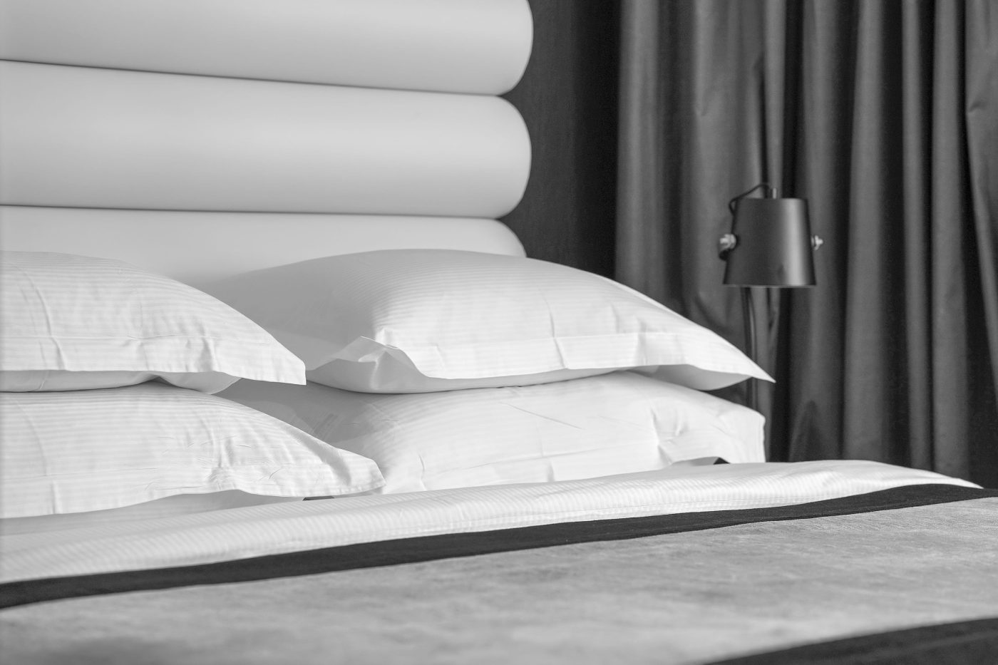 782742 Bugaderia Garraf | PymesAutonomos.com Hotel-style bed with white pillows, padded headboard, and a bedside lamp to the right with dark curtains in the background | PymesAutonomos.com